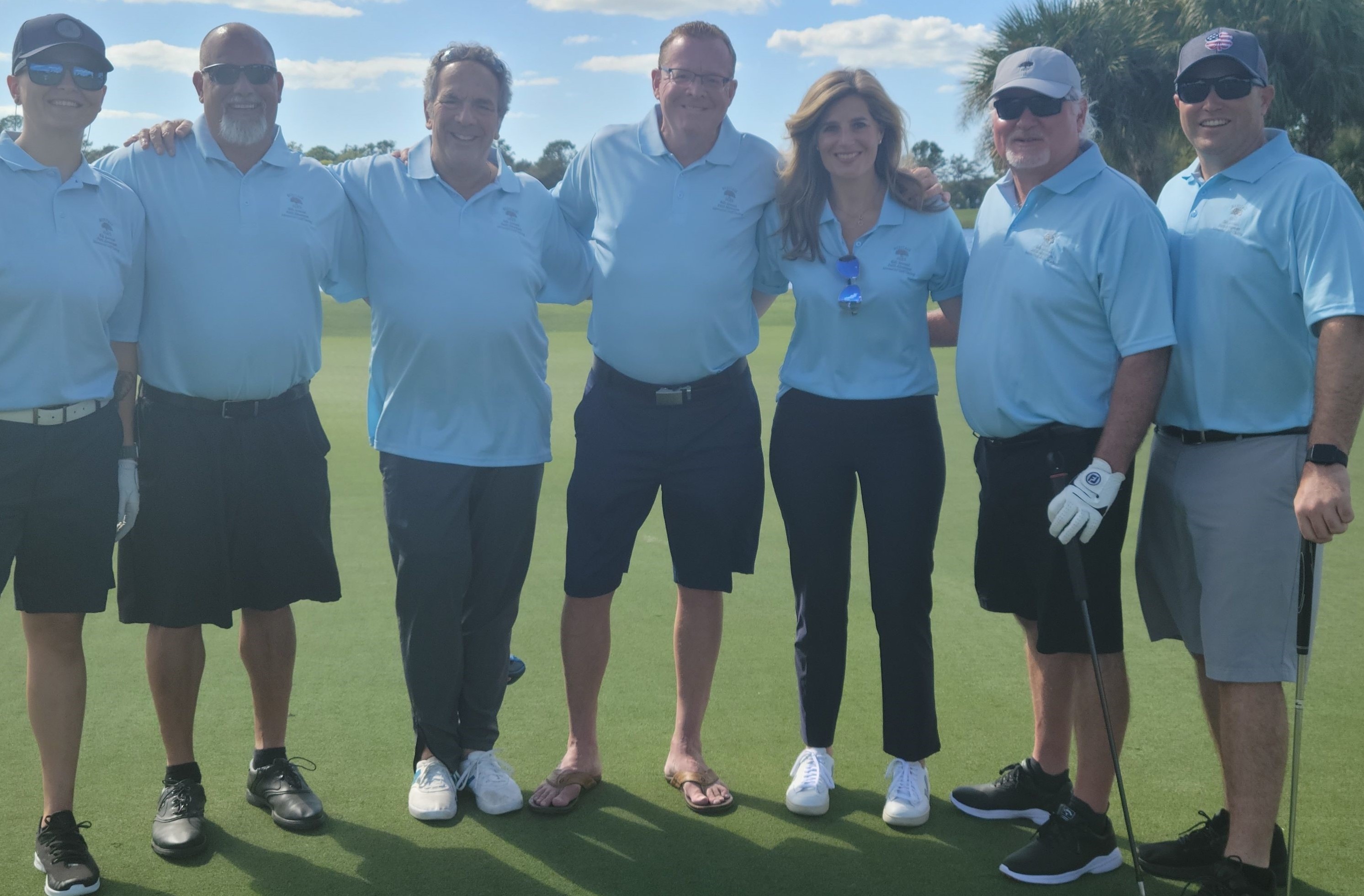 A group of seven people in pale blue golf shirts stands on a golf course